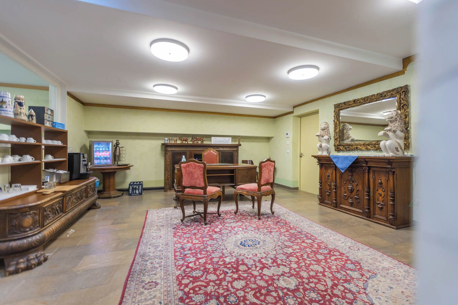 A spacious living room with a large red and white patterned rug, a wooden coffee table, two chairs, a television, and a decorative cabinet with a mirror.