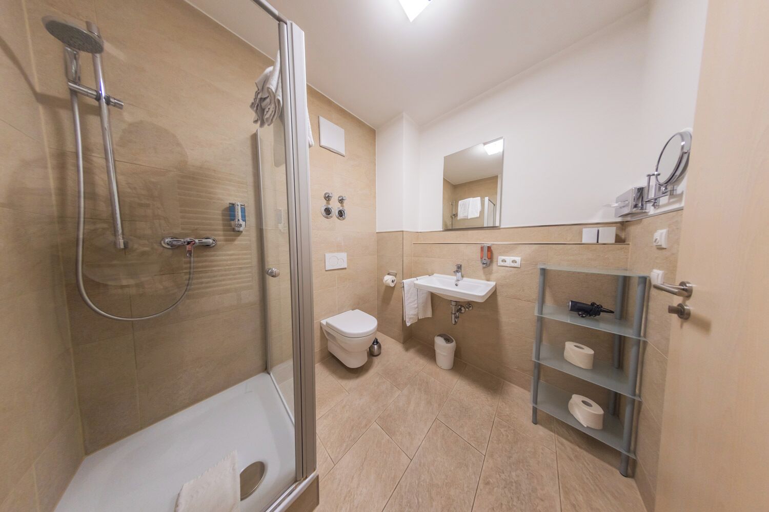 A modern bathroom with a glass-enclosed shower, a white toilet, a sink with a mirror, and a shelf with toiletries.