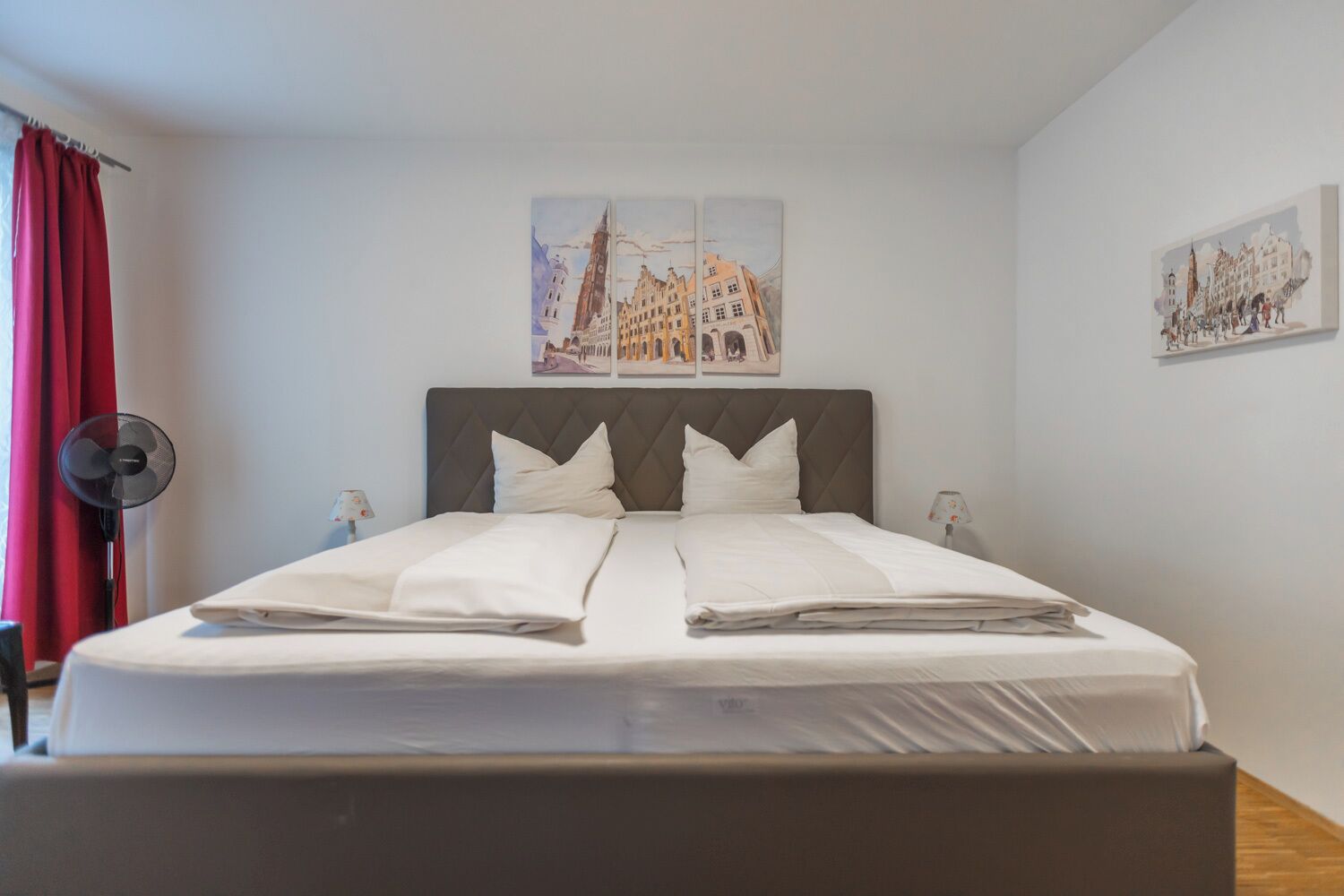 Ein ordentlich bezogenes Bett mit grauem Kopfteil, weißer Bettwäsche und zwei an der Wand befestigten Bildern mit Stadtansichten.