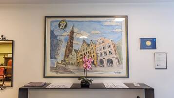 A framed painting of a European cityscape with a tall tower, a flower vase, and a framed certificate on the wall.