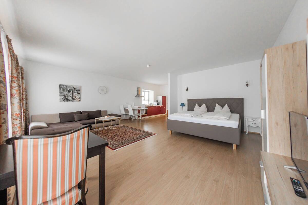 A modern hotel room with a large bed, a sofa, a dining area, and a kitchen in the background.