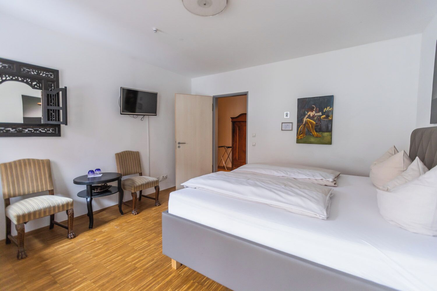 A hotel room with a bed, a TV, a desk with chairs, and a door leading to another room.