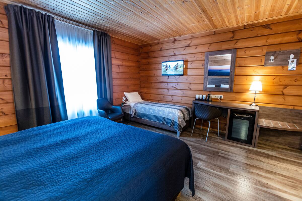 A wooden cabin room with a bed, a desk, a chair, and a small table.