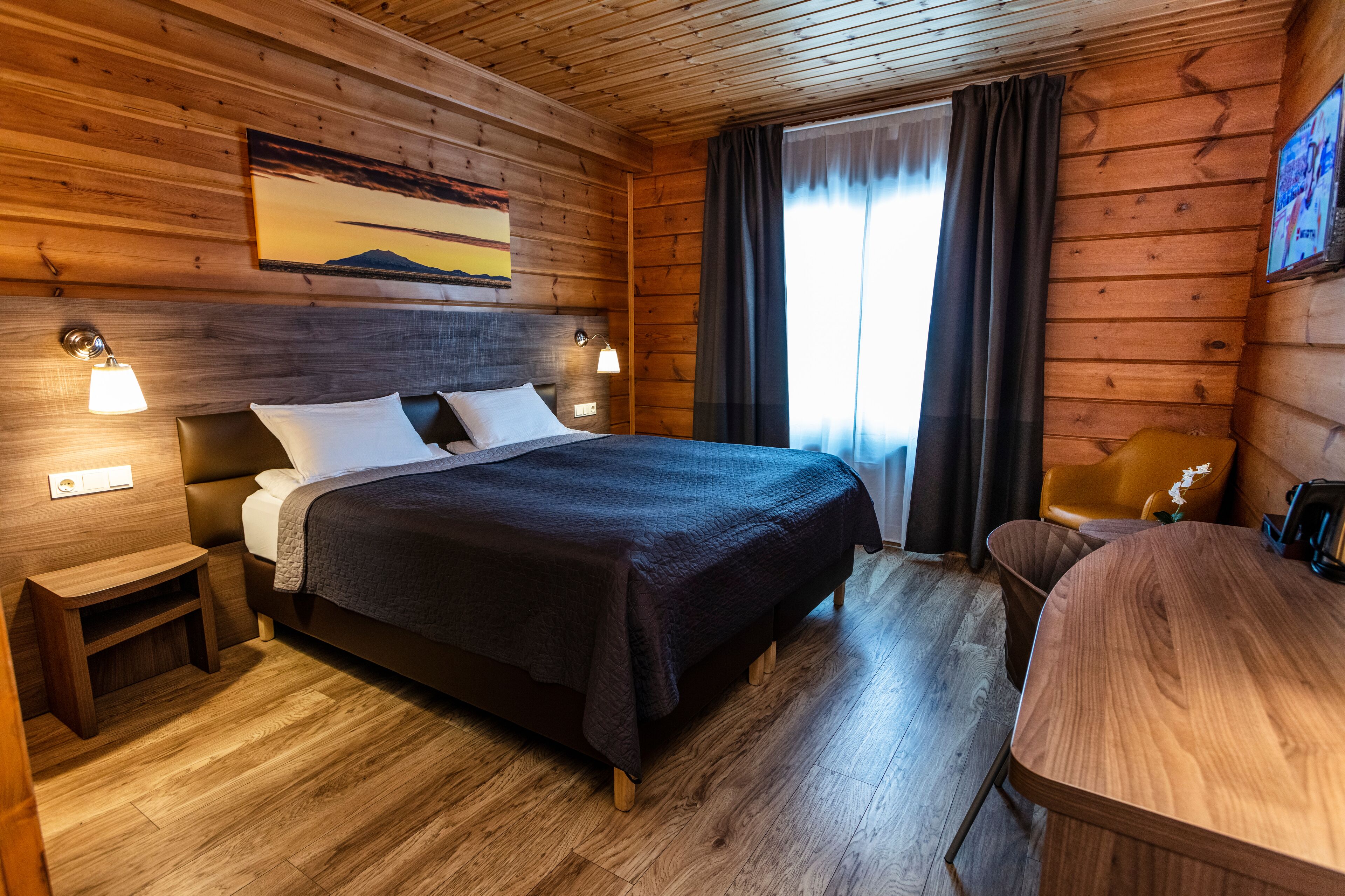 A wooden cabin room with a bed, bedside tables, a chair, and a wooden desk.