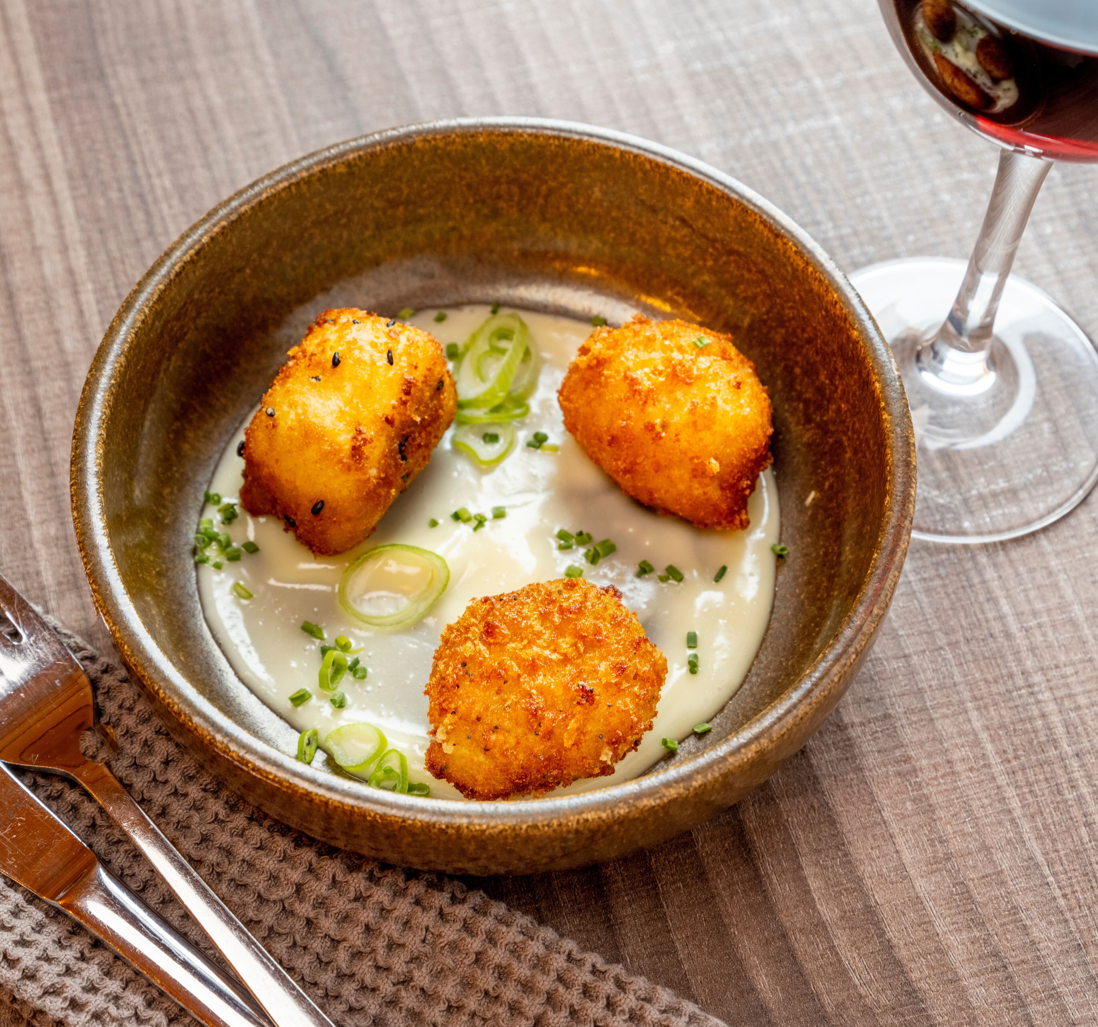 A bowl of soup with three round, breaded items and green onions, served in a ceramic bowl on a textured tablecloth.