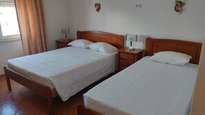 A hotel room with two single beds, wooden headboards, white linens, a nightstand with a lamp, and a window with curtains. - Alojamentos S.José (Peniche)