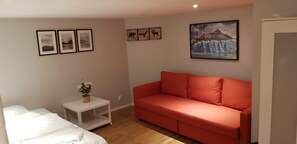 Quarto com sofá vermelho, cama branca e pequena mesa com uma planta. Quadros emoldurados na parede e uma grande imagem de montanha.