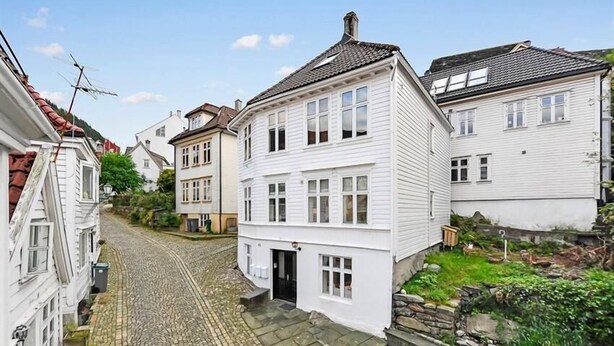 A narrow cobblestone street lined with traditional white buildings, featuring a central two-story house with a black front door and a small garden area. - Skuteviken Apartments (Bergen)