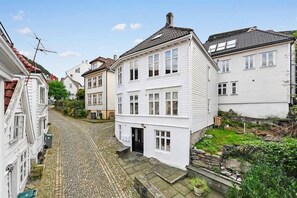 A narrow cobblestone street lined with traditional white buildings, featuring a central two-story house with a black front door and a small garden area. - Skuteviken Apartments (Bergen)
