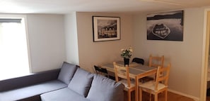 Uma sala de estar com um sofá cinzento, uma mesa de jantar de madeira com quatro cadeiras e duas fotografias emolduradas na parede.