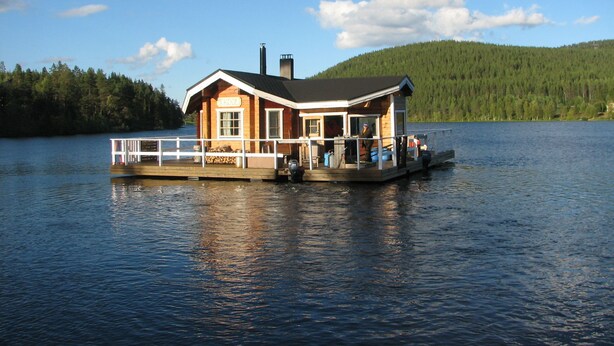 A wooden houseboat with a deck and a chimney, situated on a calm lake with a forested hill in the background. - Immelkartano (Kittila)