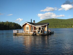 A wooden houseboat with a deck and a chimney, situated on a calm lake with a forested hill in the background. - Immelkartano (Kittila)