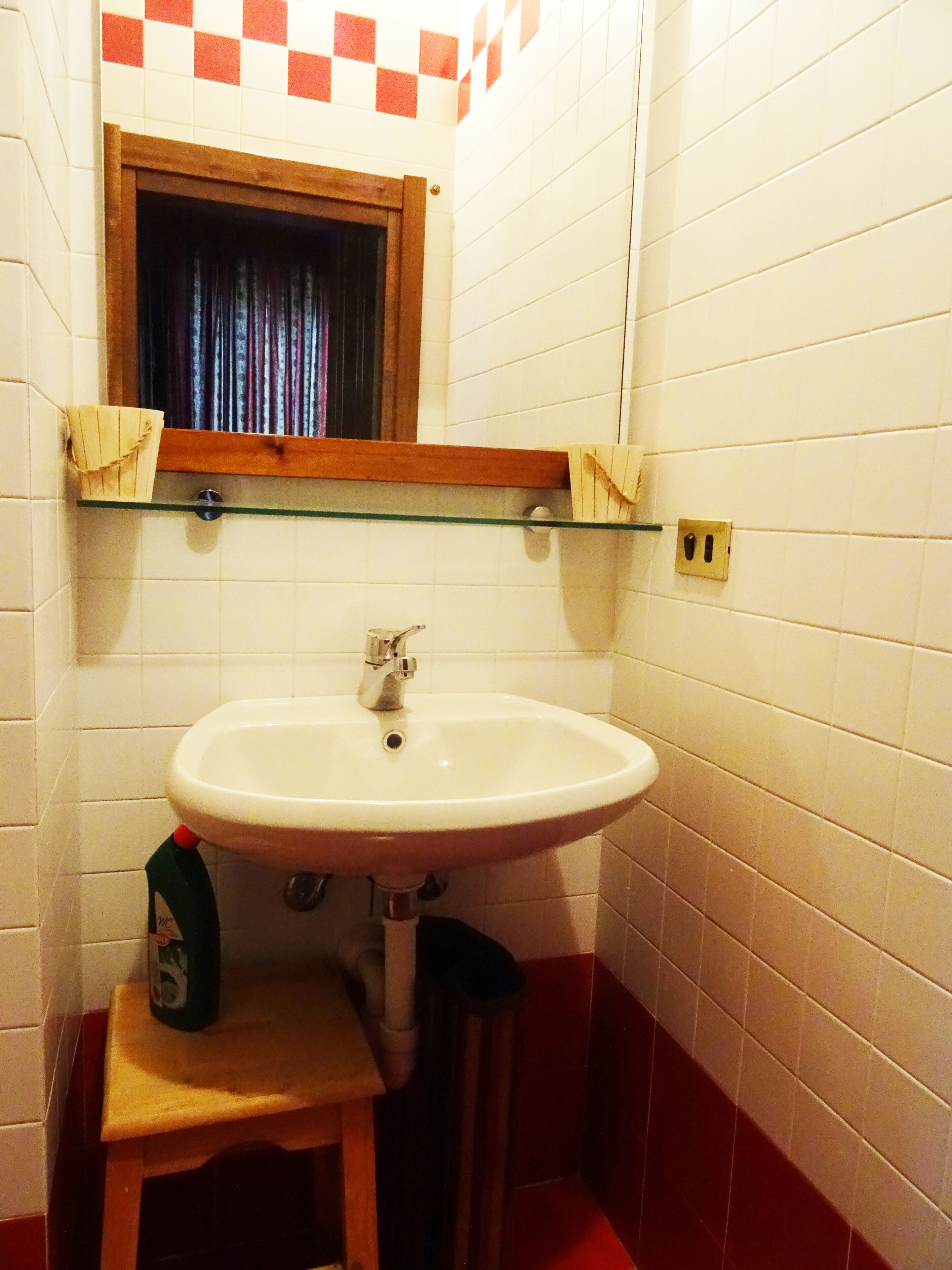A bathroom with a white sink, a wooden stool, a glass shelf above the sink, and a window with curtains.