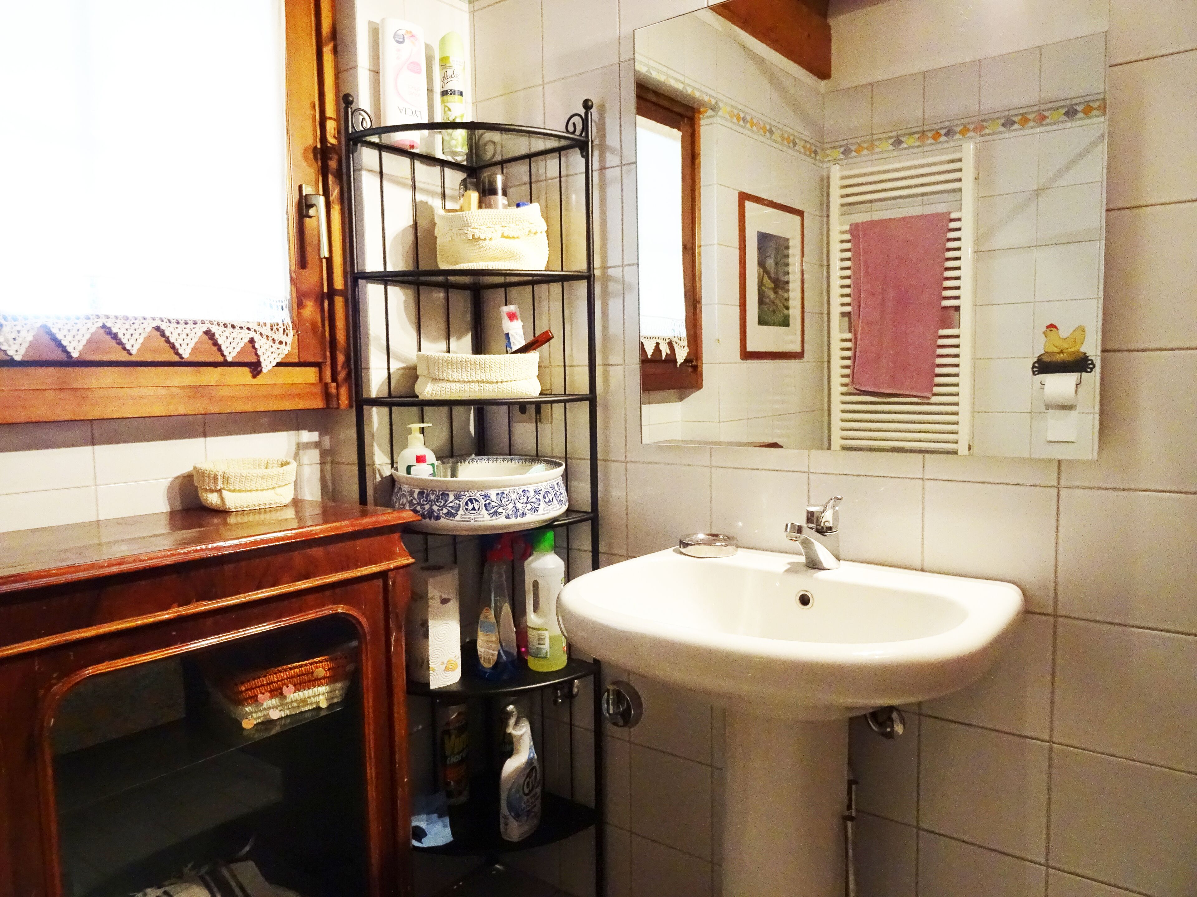 Un baño con lavamanos blanco, espejo, toallero y estantes con artículos de aseo.
