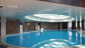 Una piscina interior con techo de tragaluces, zona acuática cristalina y sillas de descanso junto a la piscina.