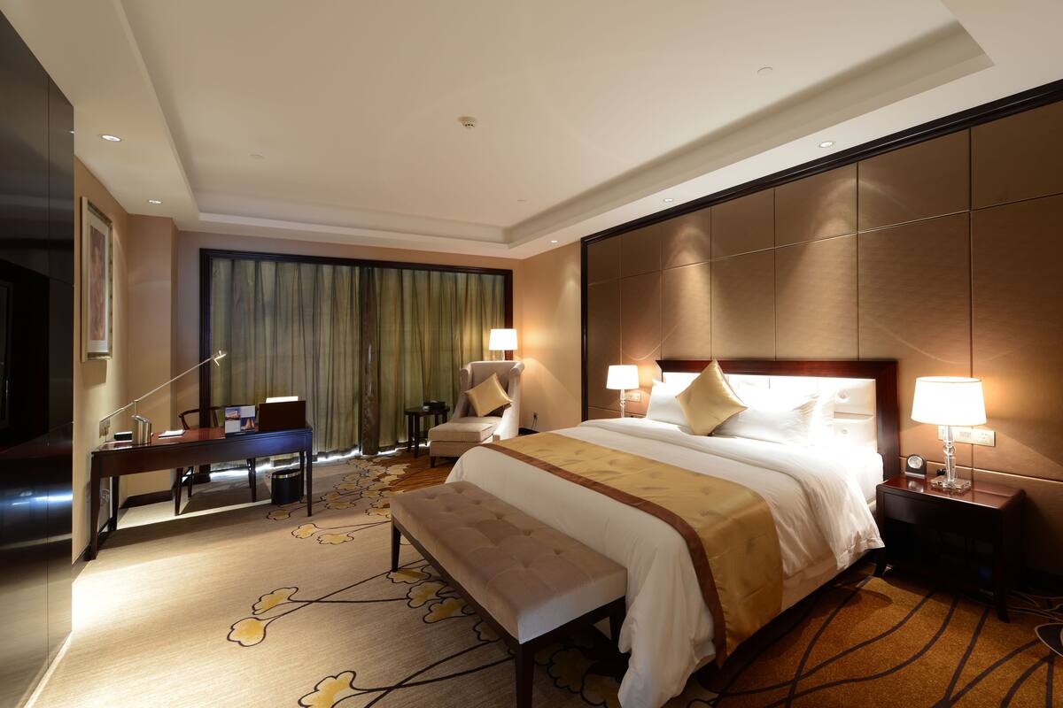 A hotel room with a large bed, a desk, a chair, a bench, and a TV.