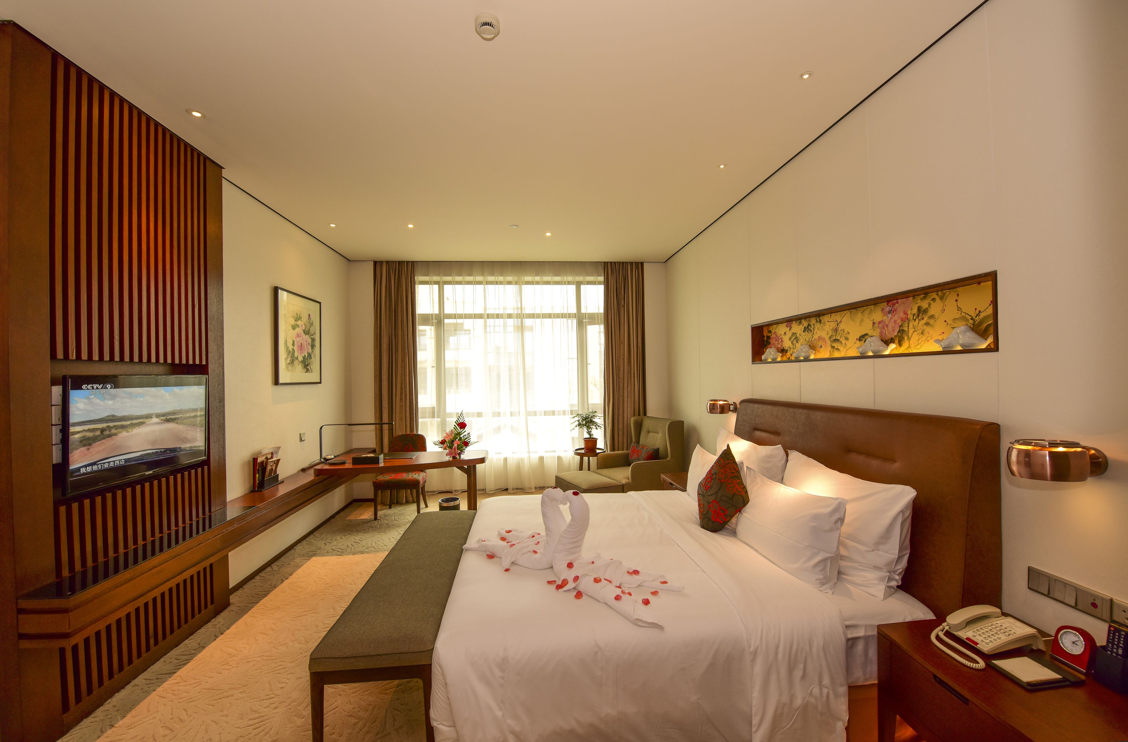 A hotel room with a large bed, a TV, a desk, a chair, a sofa, a bedside table with a lamp, and a painting on the wall.
