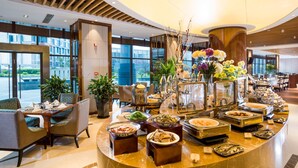 Disposição de buffet num lobby moderno de hotel, com diversas opções de comida, áreas de estar e grandes janelas que oferecem vista para o exterior.