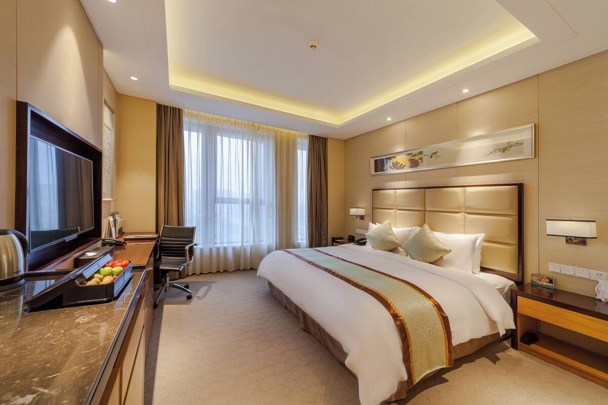 Ein Hotelzimmer mit einem großen Bett, einem Flachbildfernseher, einem Schreibtisch mit Obstschale und einem Fenster mit Vorhängen.