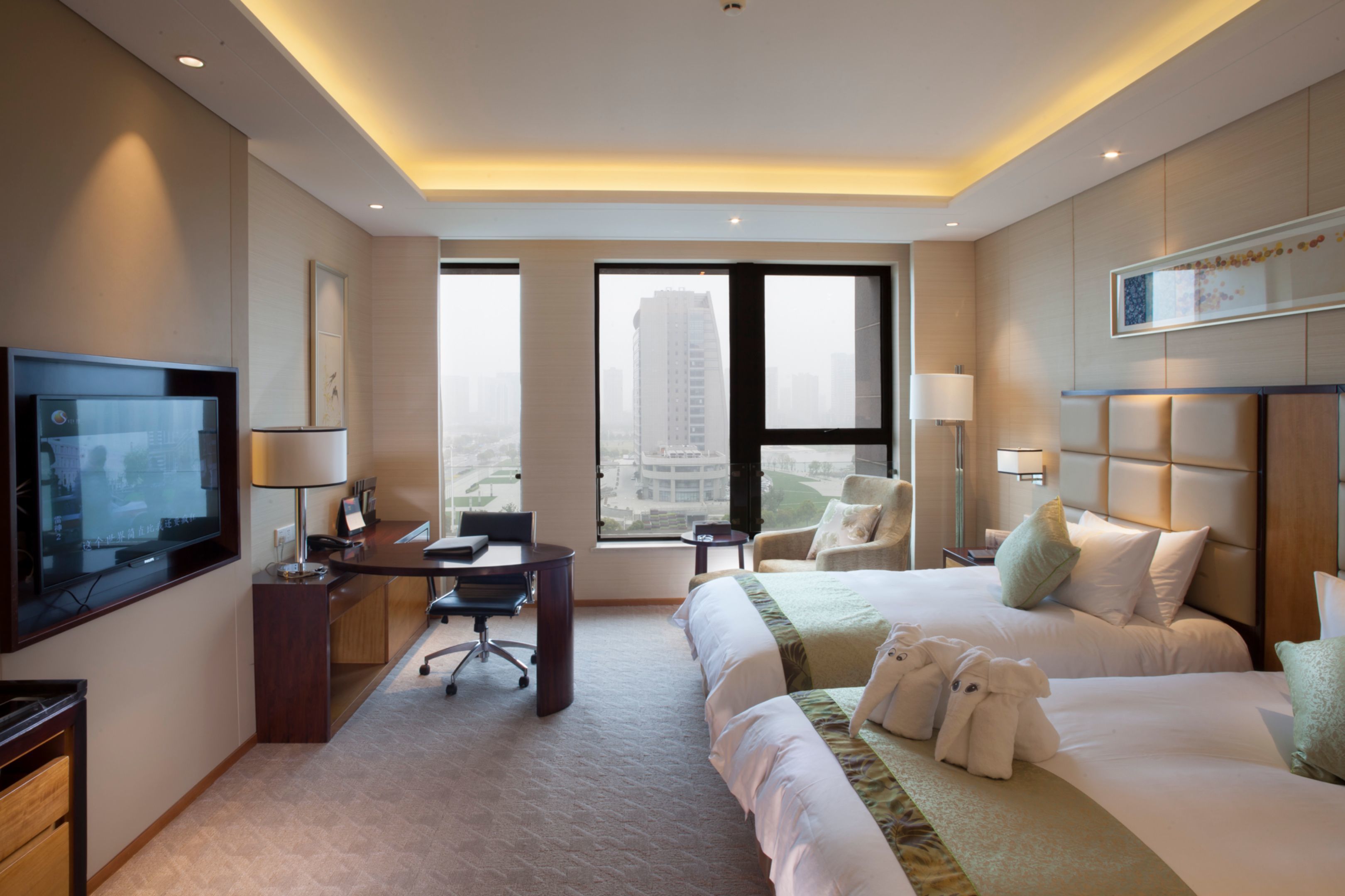 A hotel room with a large bed, a desk, a chair, a TV, and a view of a cityscape through large windows.