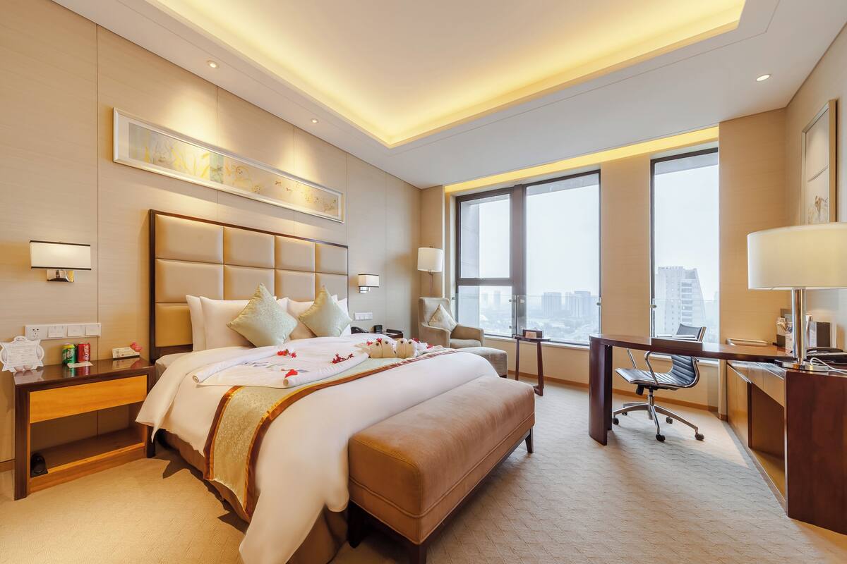 Ein Hotelzimmer mit einem großen Bett, Nachttischen, einem Schreibtisch und einem Sessel. Große Fenster bieten Blick auf die Stadt.