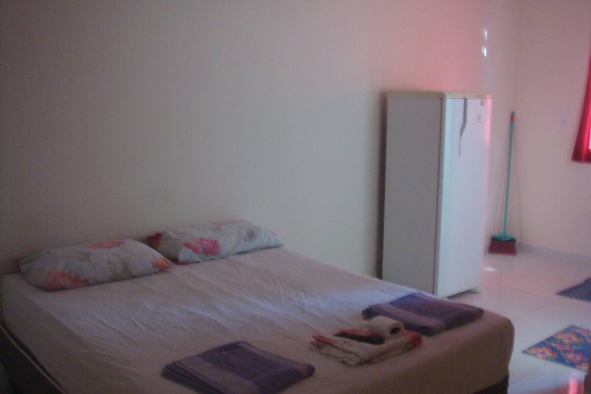 Ein Einzelbett mit weißer Matratze, zwei Kissen und einer gefalteten Decke. Ein kleiner Tisch steht neben dem Bett mit ein paar Gegenständen darauf.