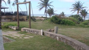 A grassy area with a stone wall and a wooden structure, palm trees, and a view of the ocean. - Stellamares Apartamentos (Salvador)