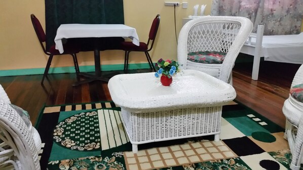 A room with a white wicker sofa, a matching ottoman, and a dining table with a white tablecloth. A red vase with flowers is on the ottoman. The room has a patterned rug and a green curtain in the background. - Lake Mainstay Resort (Anna Regina)
