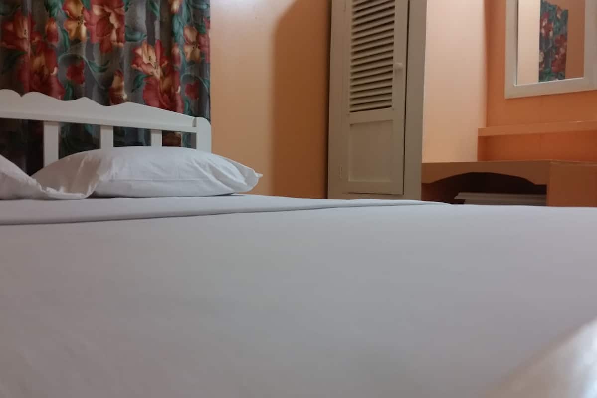 Ein Bett mit weißer Bettwäsche und Kissen, ein weißer Kopfteil, ein Fenster mit blumigen Vorhängen, eine geschlossene Tür und ein gerahmtes Bild an der Wand.