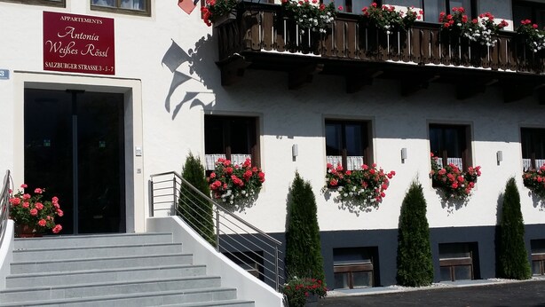 Ein Gebäude mit einem Schild für 'Antonia Weifes Rössl Apartments' im Erdgeschoss. Vor dem Eingang führen Treppen hinauf, und Blumenkästen mit roten Blumen befinden sich an den Fensterbänken.