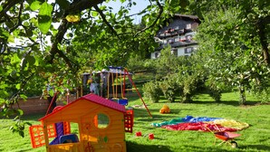 Ein Spielbereich für Kinder mit einem kleinen Haus und Schaukeln, umgeben von Grünflächen und einem Haus im Hintergrund.