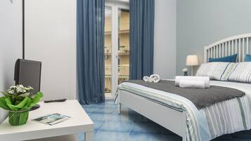 A neatly arranged hotel room with a bed, a desk with a computer and a plant, and a balcony visible through the curtains.
