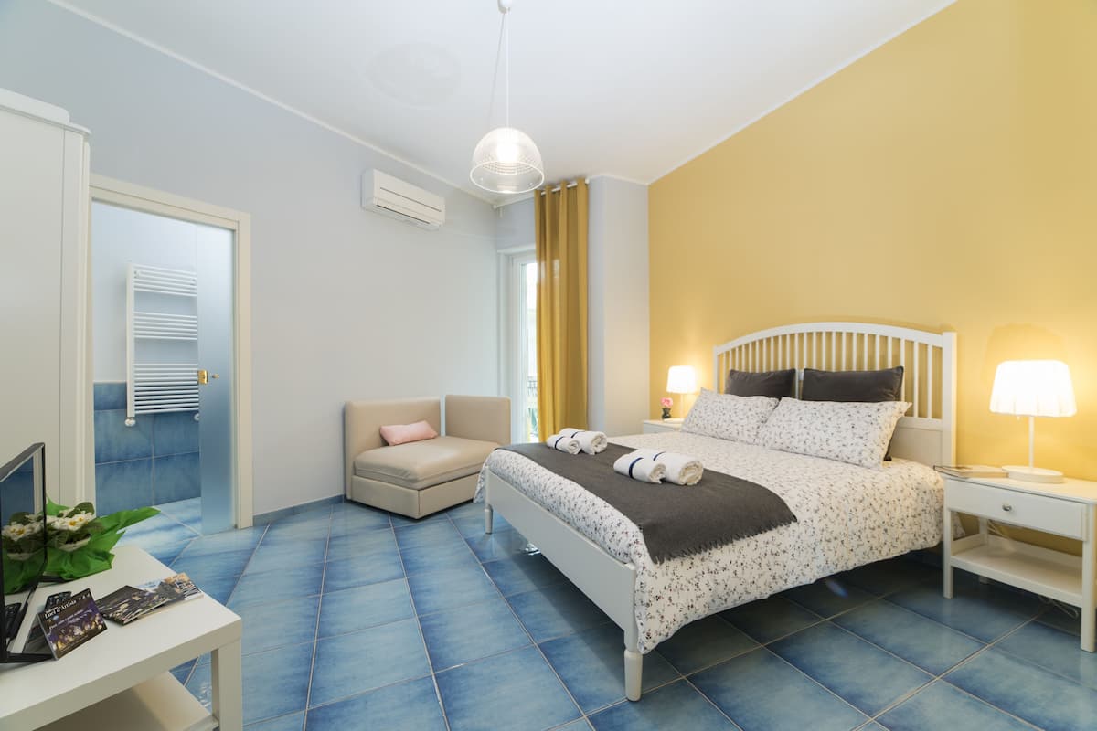 Ein Schlafzimmer mit einem Bett, einem Nachttisch, einem Sessel, einem Schreibtisch mit Computer und Büchern sowie einem Fenster mit Vorhängen.