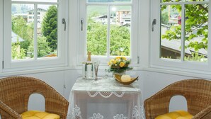 Una habitación con vistas a zonas verdes y edificios, una mesa con mantel de encaje, dos sillas de mimbre con cojines amarillos y un jarrón con flores.