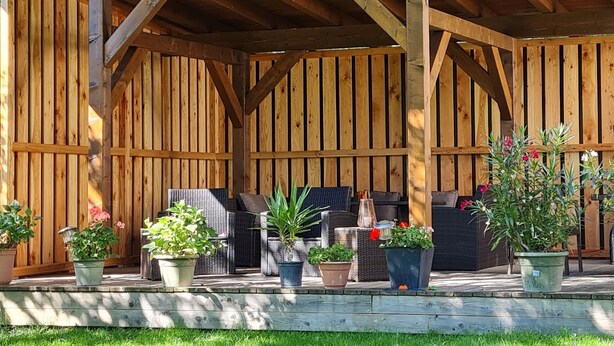 Un patio techado con vigas y cerca de madera. Hay varias plantas en macetas y mobiliario exterior, incluyendo una mesa y sillas.