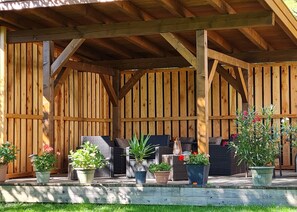 Una veranda riparata con travi e recinzione in legno. Diverse piante in vaso e arredi da esterno, tra cui un tavolo e sedie.