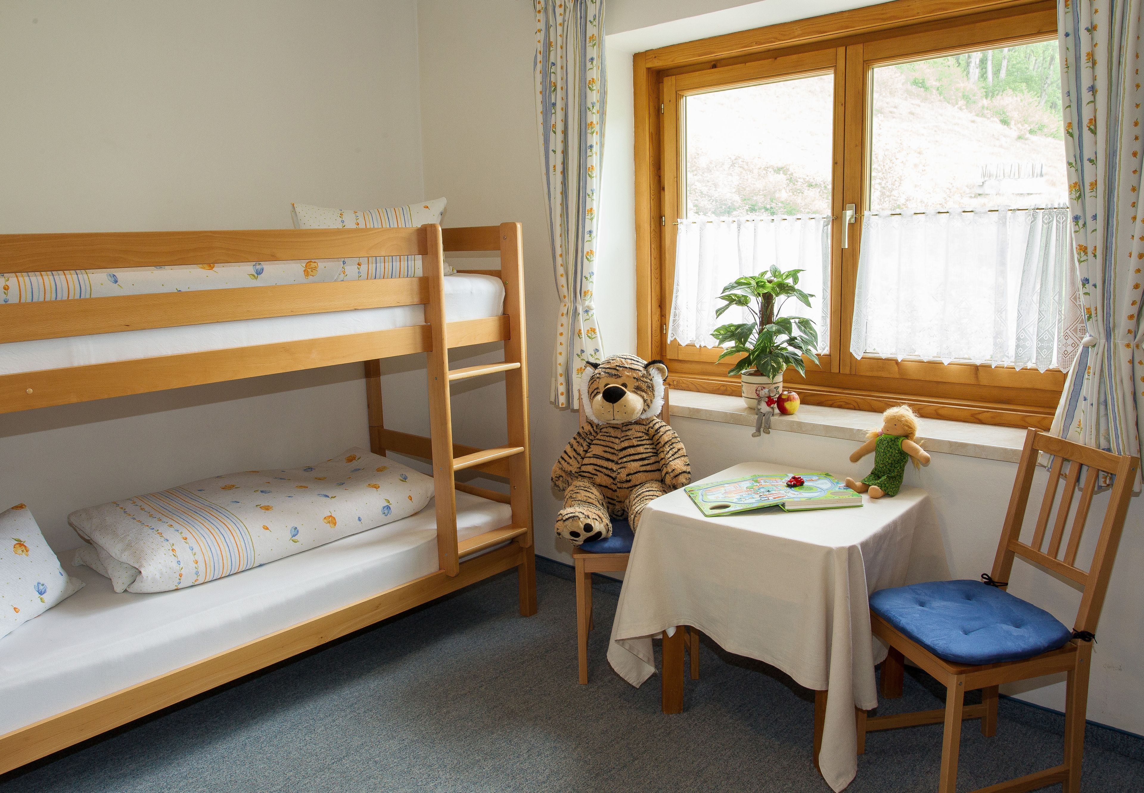 Una habitación infantil con literas, un escritorio, una silla, un osito de peluche y una ventana con cortinas.