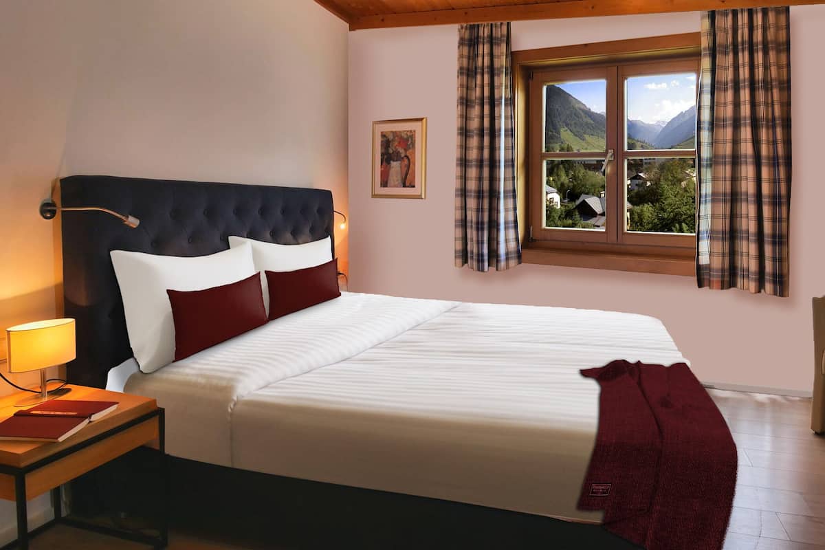 A bedroom with a large bed, a nightstand, a lamp, a window with curtains, and a painting on the wall.