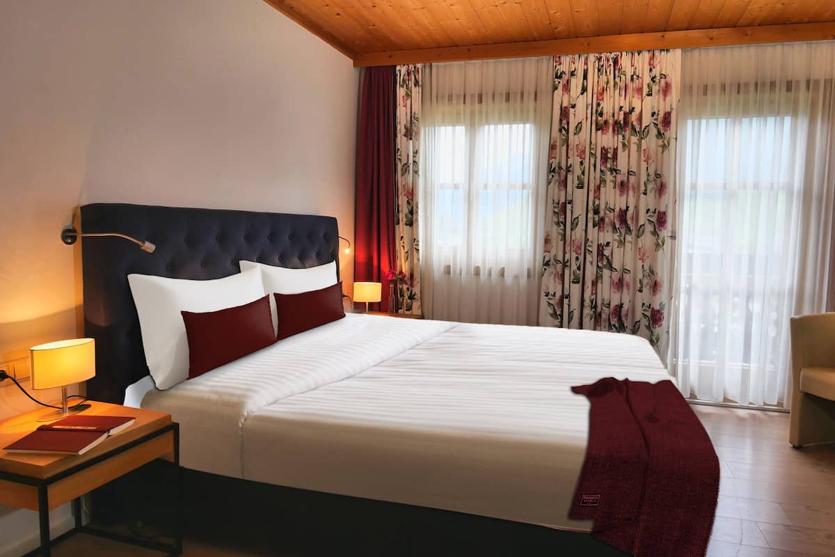 A hotel room with a large bed, a wooden headboard, a nightstand with a lamp, a chair, and floral curtains.