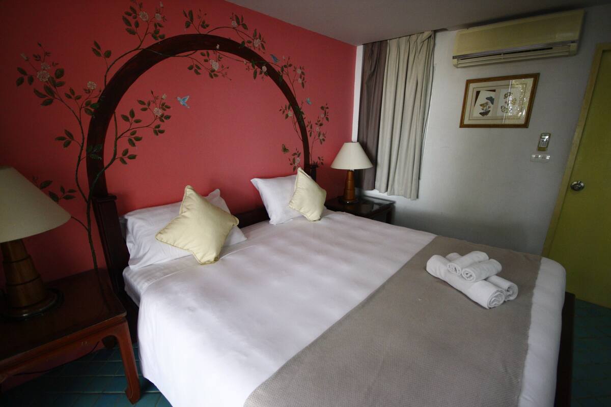 A neatly made bed with white linens and a beige throw, a wooden nightstand with a lamp, and a floral wall mural.
