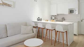 A modern kitchen with white cabinets, a microwave, and a sink. There is a white bar stool and a small round table in front of a white sofa.
