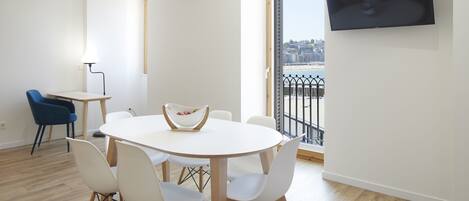 A modern living room with a round white table, white chairs, a blue chair, a TV on the wall, and a balcony with a view.