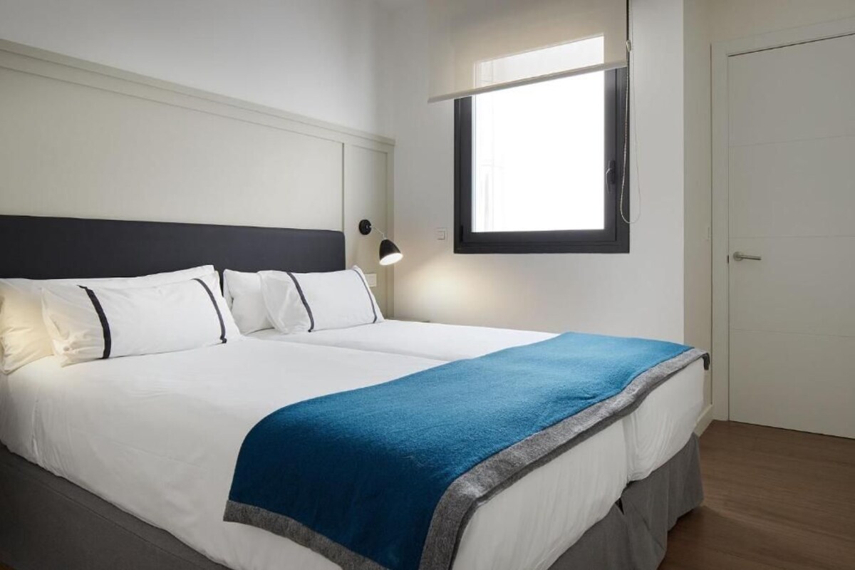 Ein ordentlich bezogenes Bett mit weißer Bettwäsche und blauem Akzent, eine Nachttischlampe und ein Fenster mit Blick nach draußen.