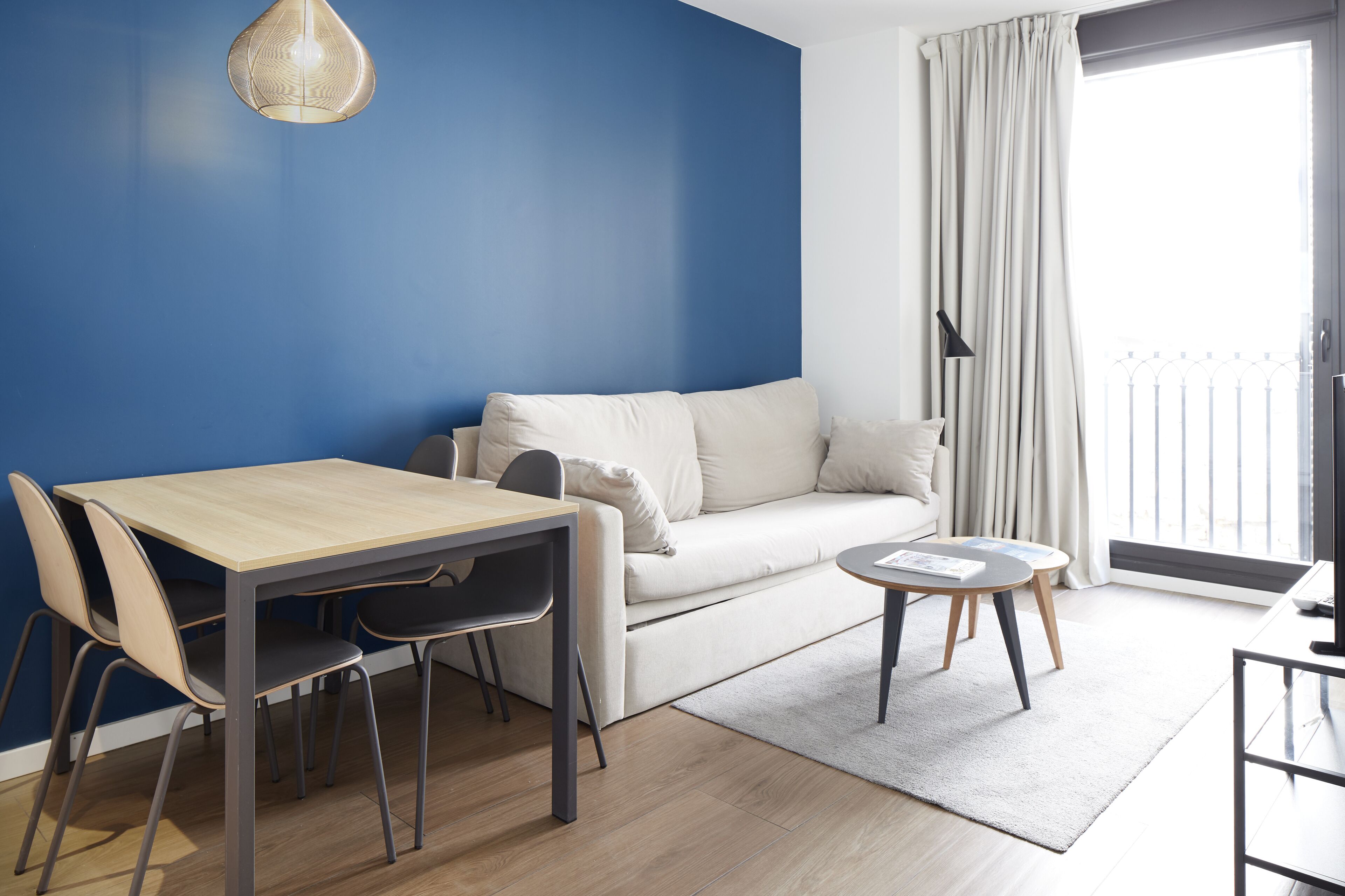 Un salon moderne avec un mur d’accent bleu, un canapé blanc, une table à manger en bois avec des chaises, et une petite table ronde avec une lampe.