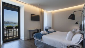 Una habitación de hotel moderna con una cama grande, un escritorio, una silla, un televisor montado en la pared y un balcón con vistas a la ciudad.