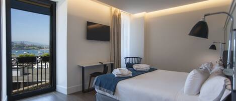 A modern hotel room with a large bed, a desk, a chair, a TV mounted on the wall, and a balcony with a view of the city.