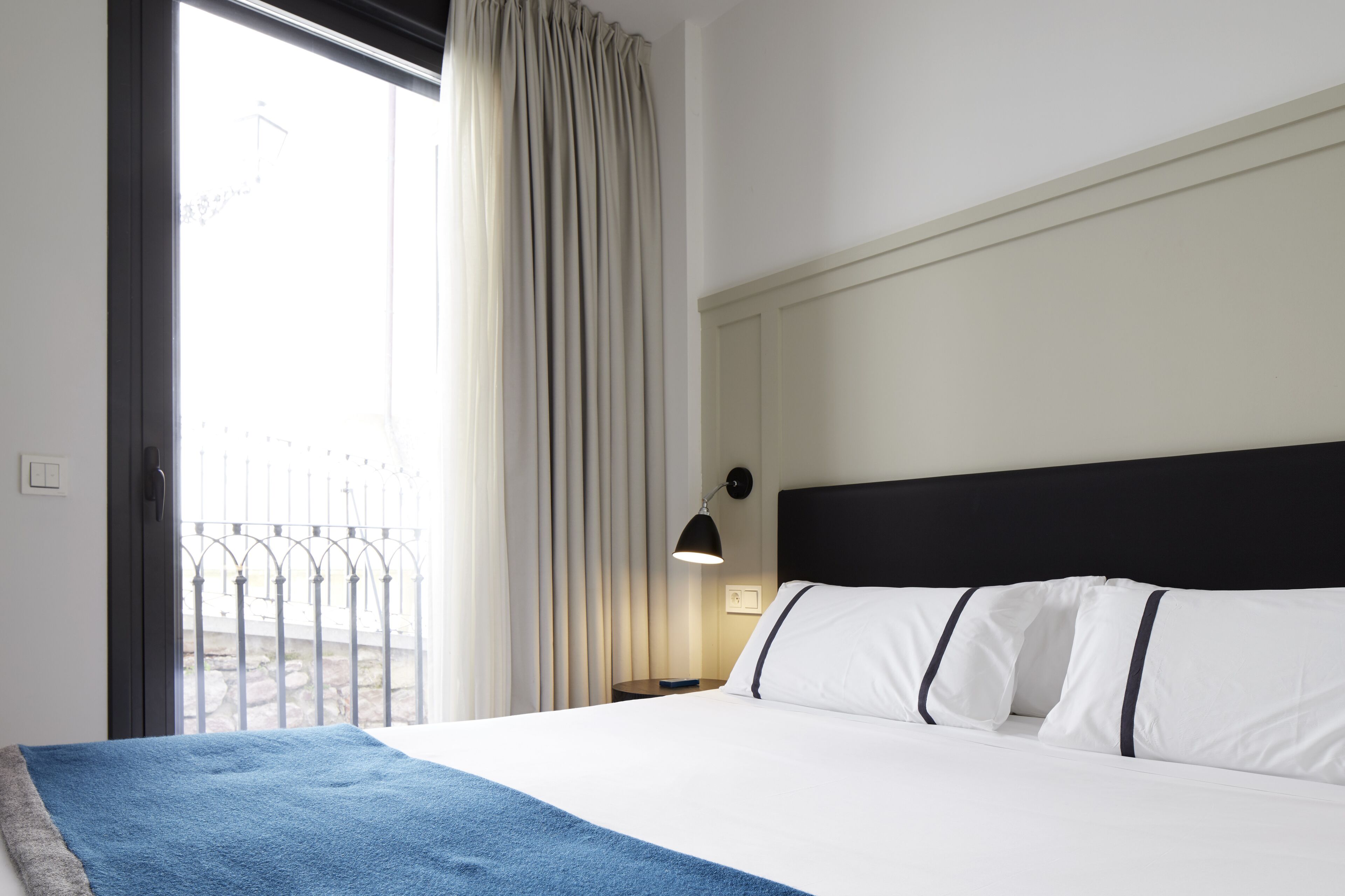 A hotel room with a bed, a nightstand, a lamp, and a view of the outside.