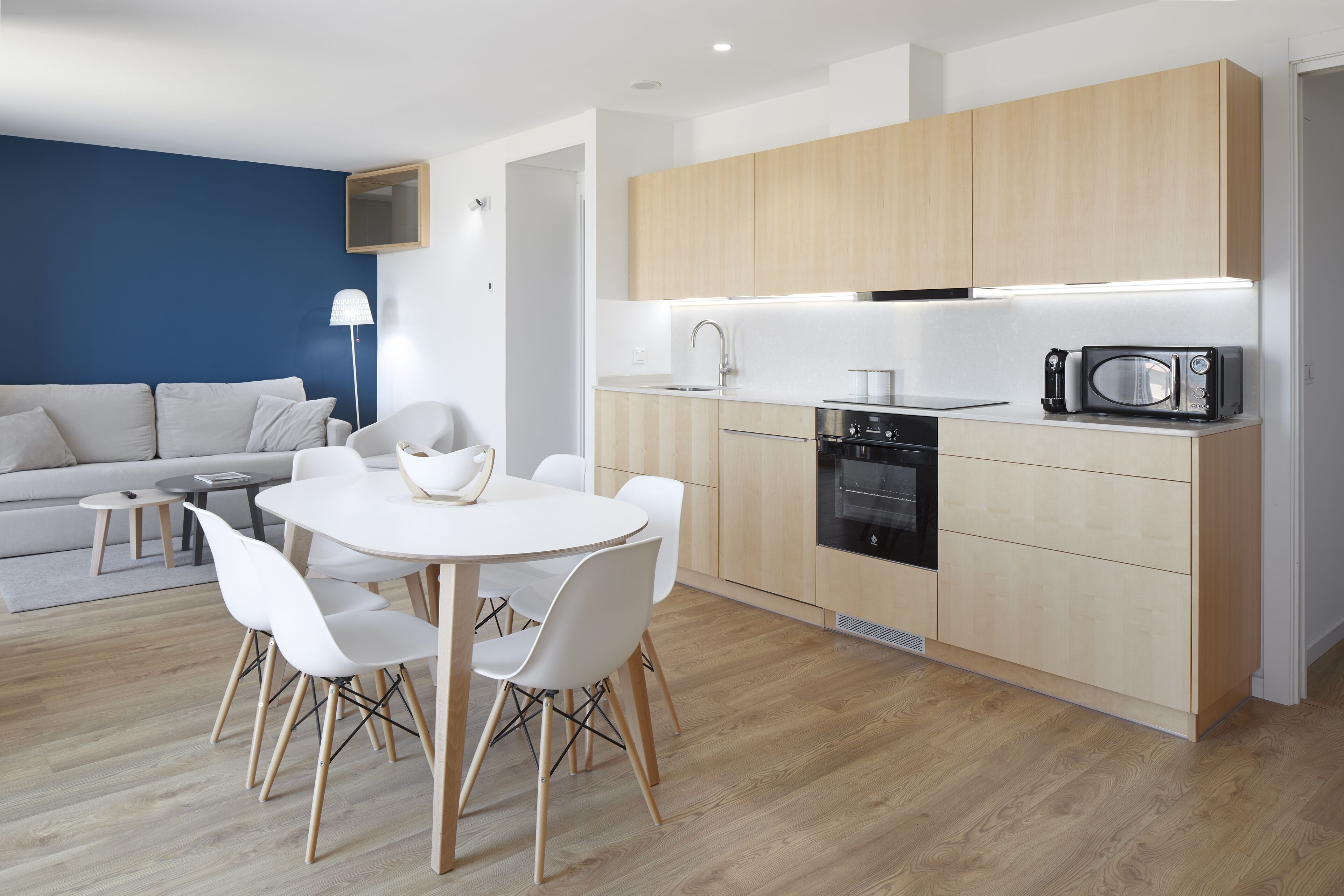 A modern kitchen with wooden cabinets, a microwave, and a stove. There is a dining area with a white table and chairs.