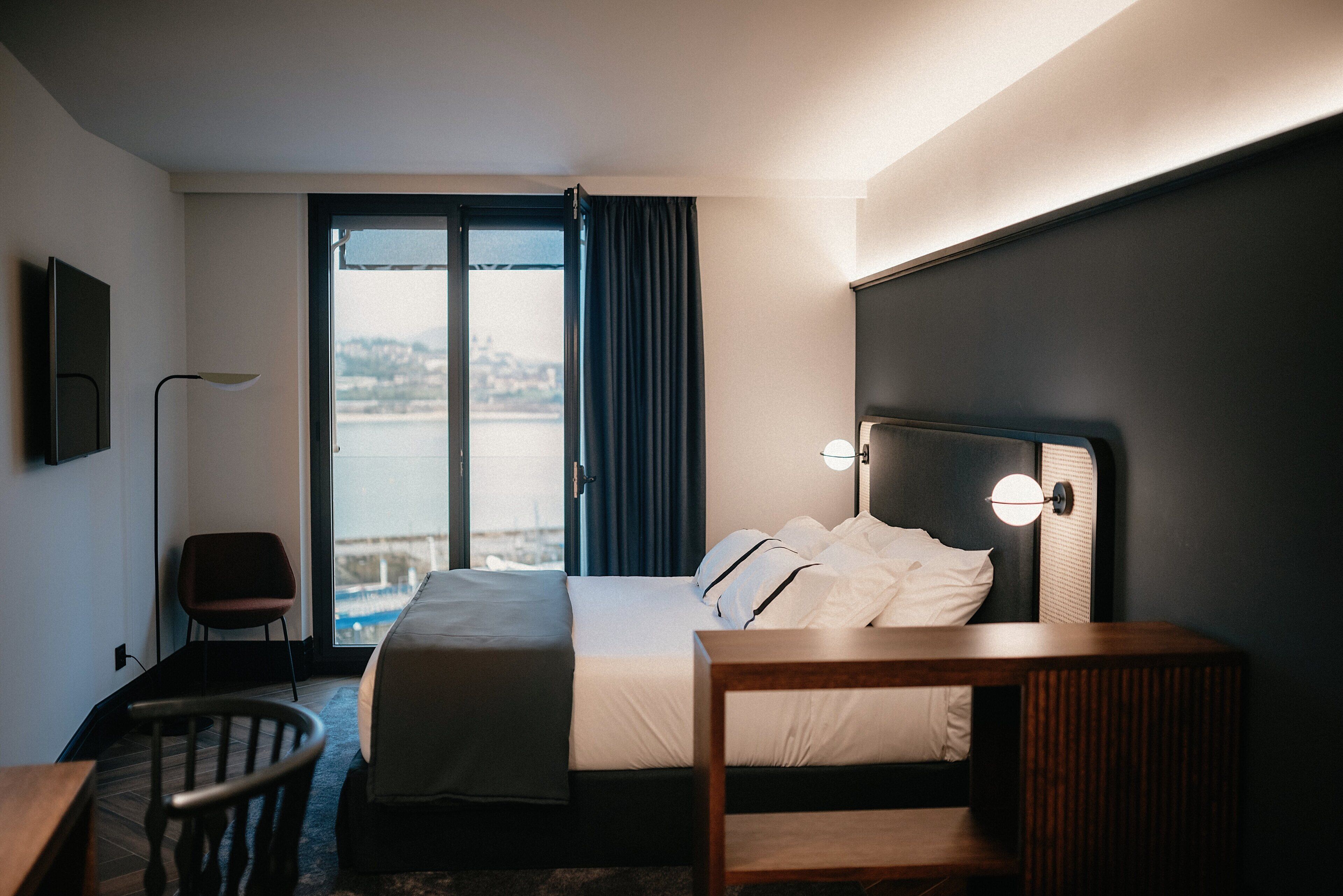 Habitación de hotel con cama, escritorio, silla y vista a un cuerpo de agua a través de un ventanal grande.