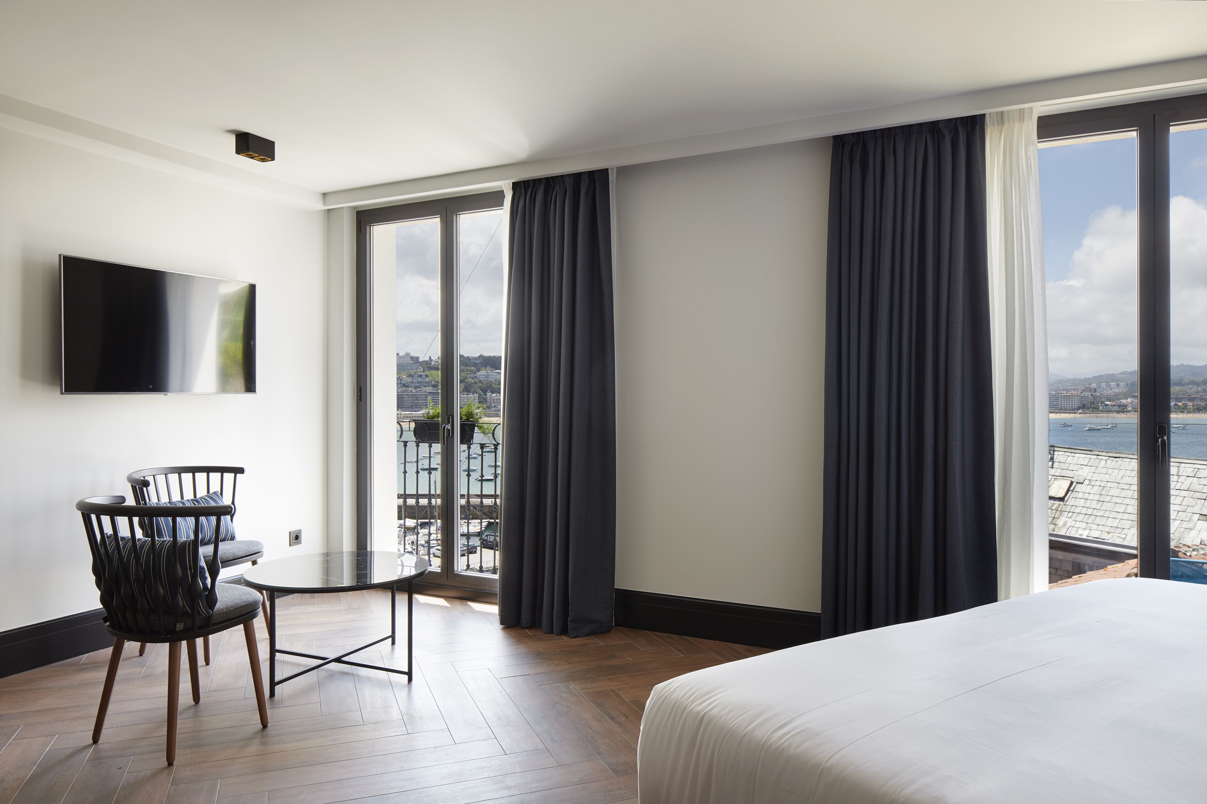 Een hotelkamer met een bed, een aan de muur bevestigde televisie, een balkon met gordijnen en uitzicht op zee.